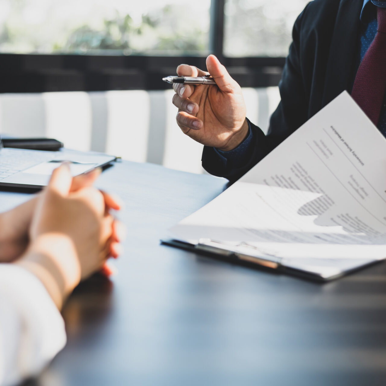 Businessman in suit in his office showing an insurance policy and pointing with a pen where the policyholder must to sign. Insurance agent presentation and consulting insurance detail to customer. Business meeting with documents and laptop.